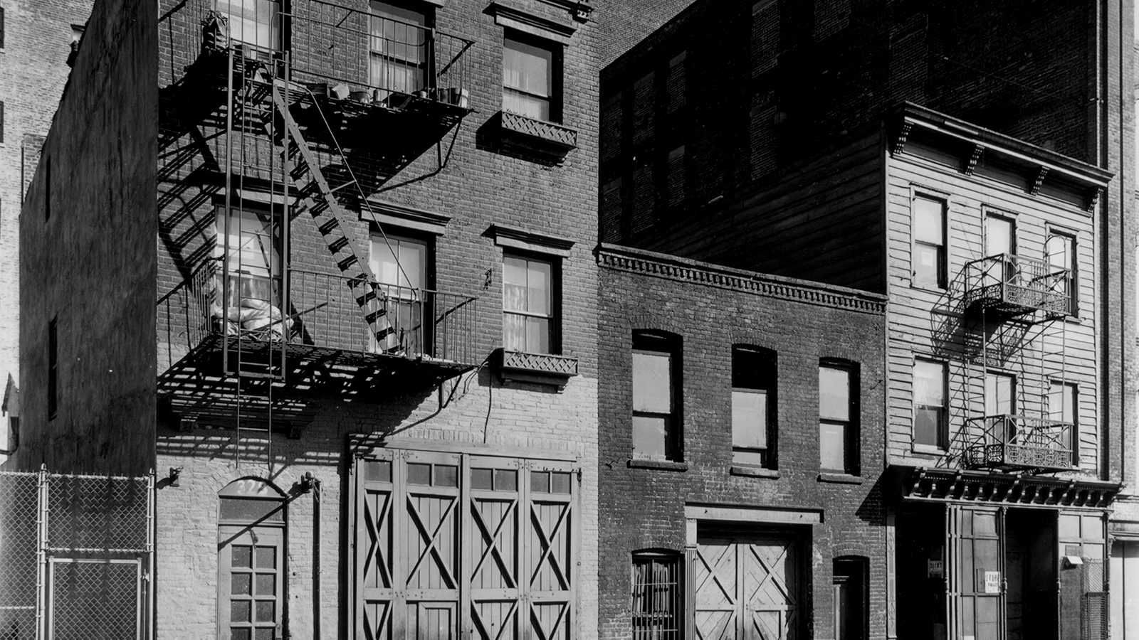 Old New York brick and wooden buildings with fire escapes, arched windows and large carriage doors in black and white
