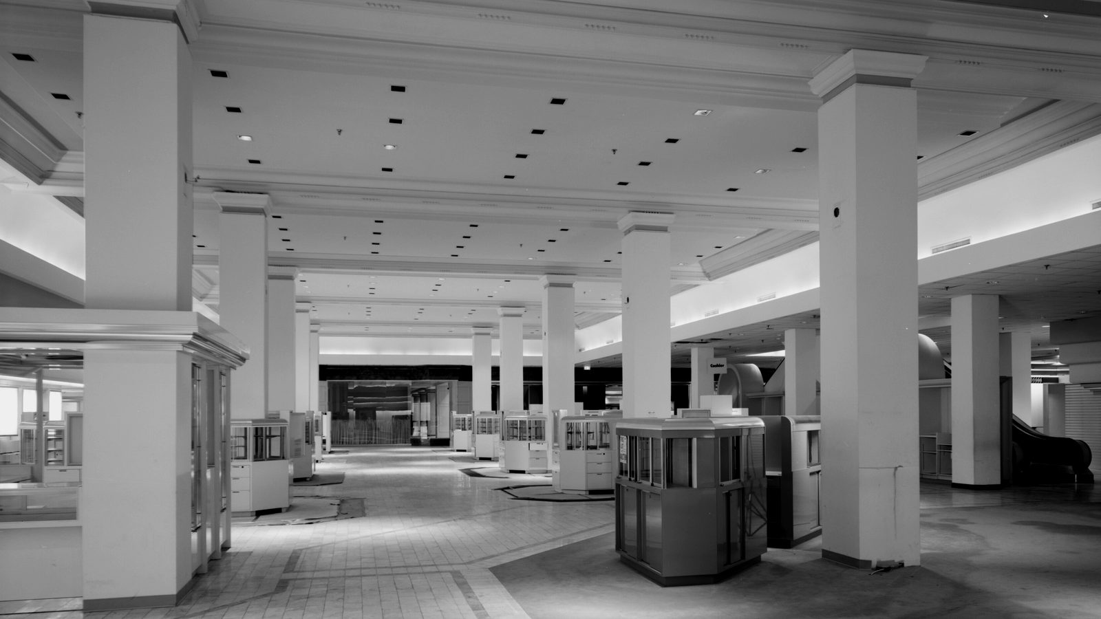 Empty department store sales floor with tall white columns, vacant display counters, a cashier kiosk and an escalator