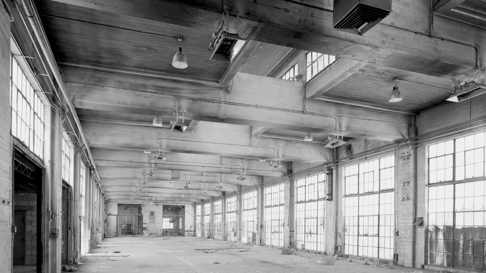 Empty factory hall with concrete beams, hanging lamps and tall steel-framed windows lining one wall
