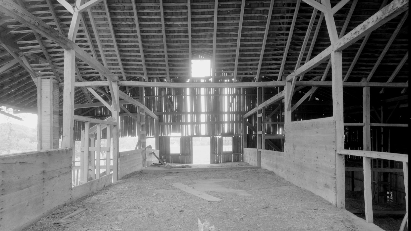 Interior of an empty timber-frame barn with wooden stalls, a straw-covered floor and light coming through the far doors