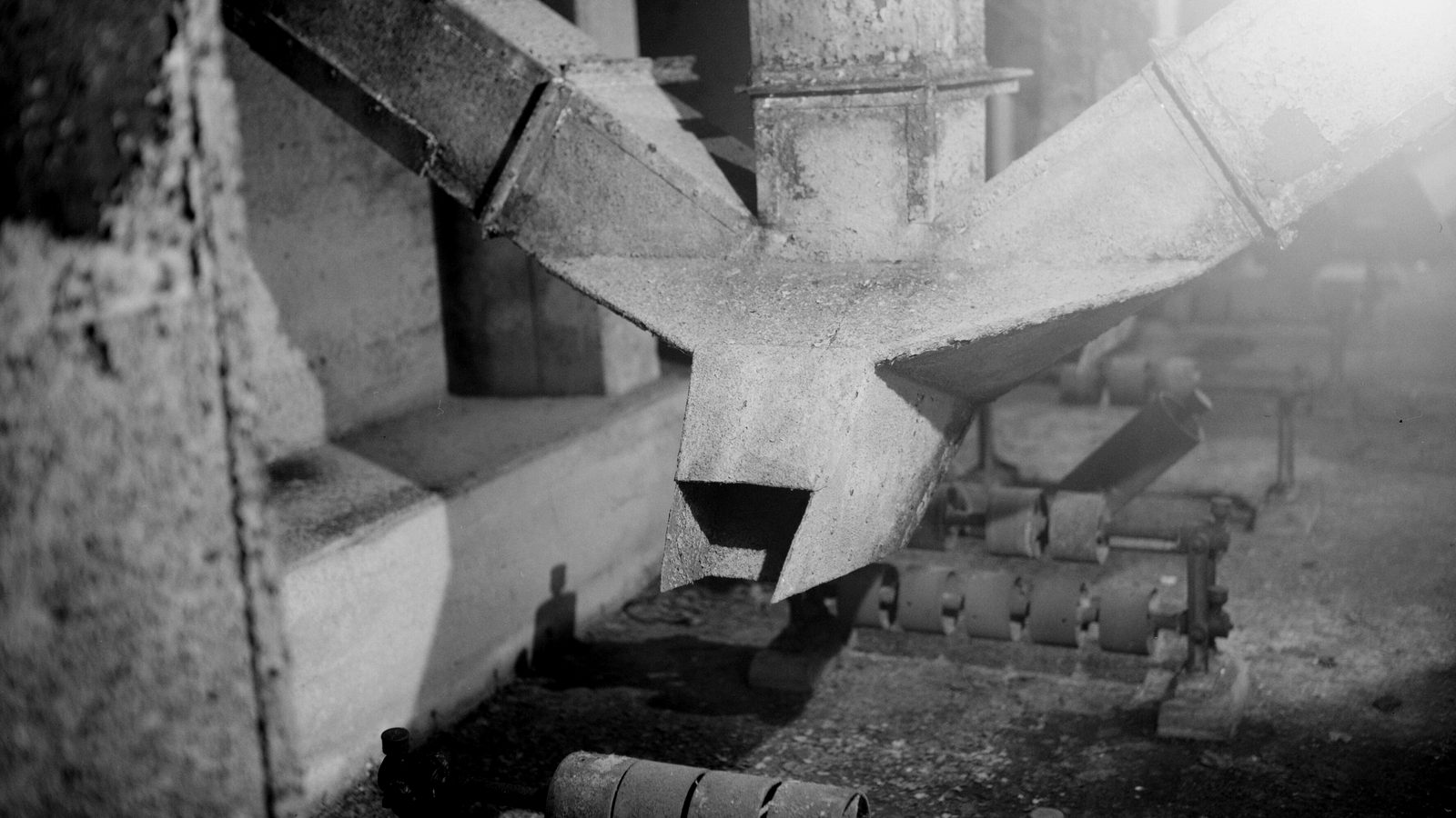 Two angled metal chutes merging into a single spout above a roller conveyor inside a factory