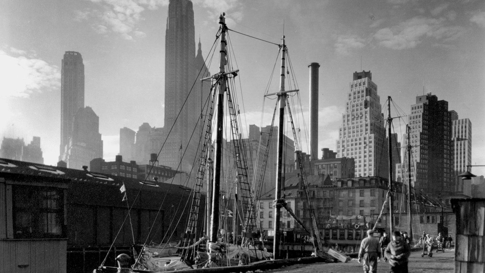 Black and white 1930s view of sailing ship masts at a Lower Manhattan dock with skyscrapers rising behind
