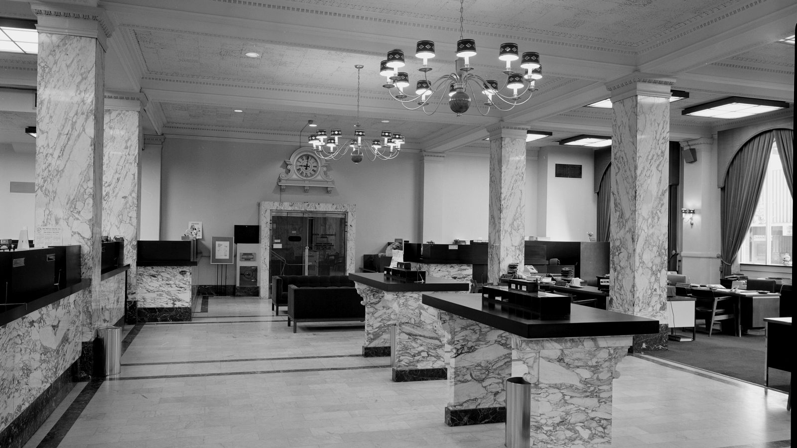 Bank lobby with marble columns, marble check-writing tables, teller counters, chandeliers and a wall clock above a vault door
