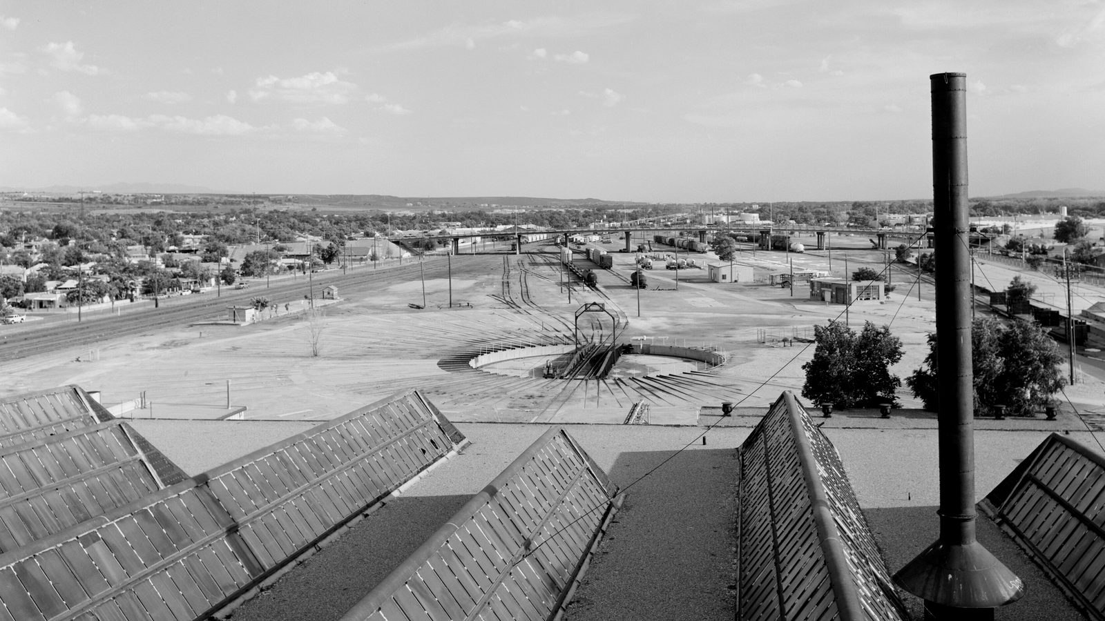 Rail yard with a turntable and freight cars seen from a roundhouse roof with skylights and a smokestack
