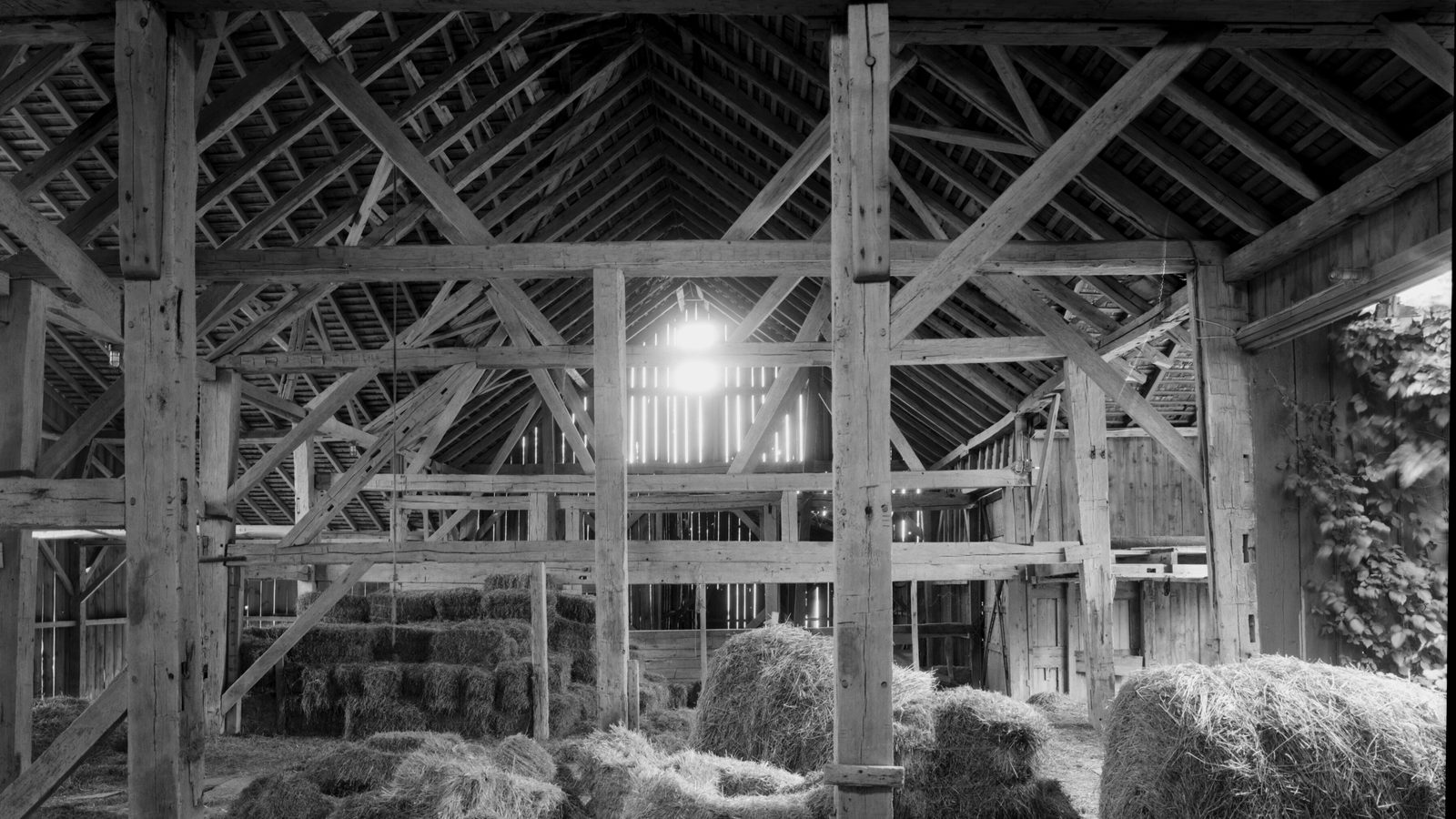 Interior of a timber-framed barn with heavy wooden posts, braced roof trusses, light through slatted boards, and stacked hay
